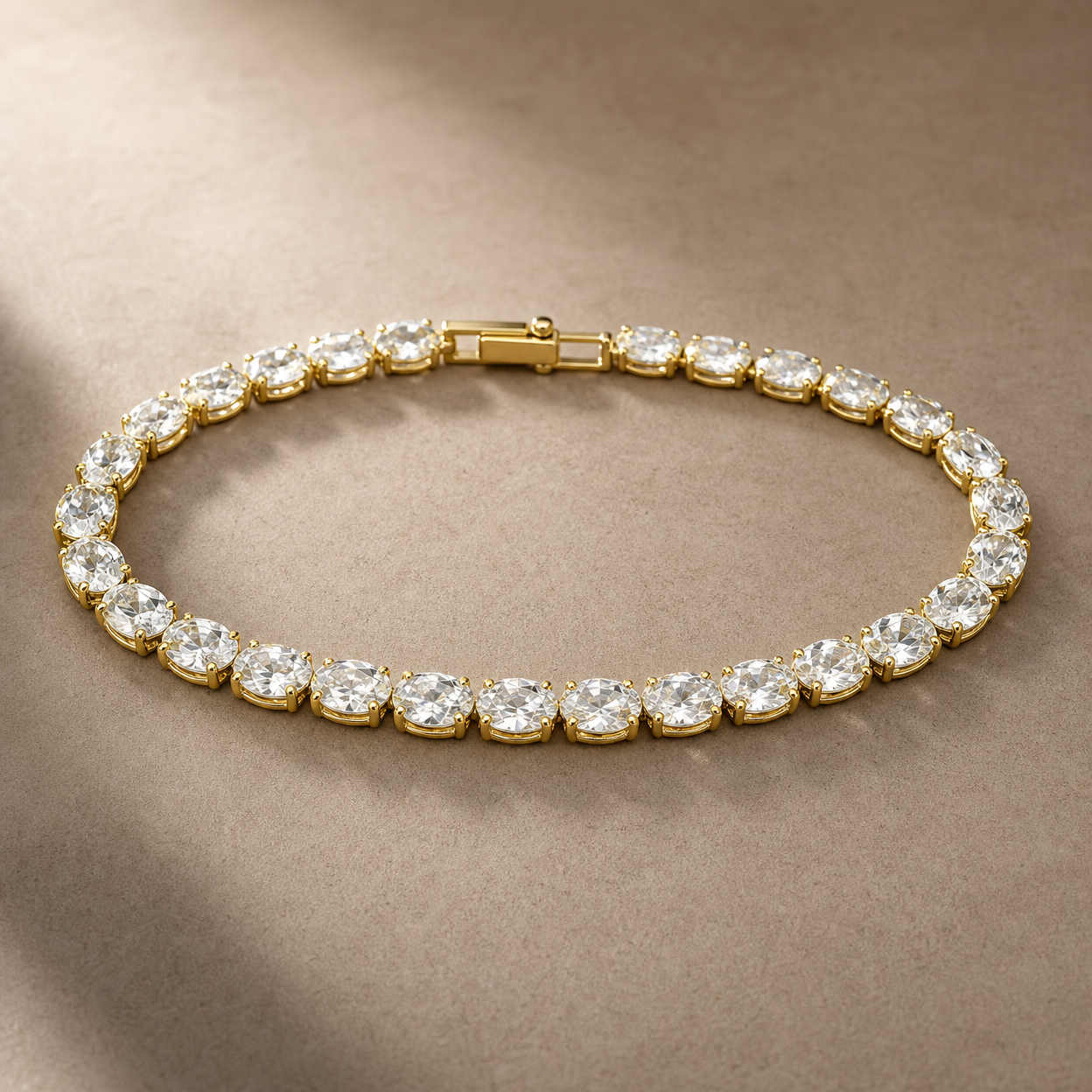 Gold-tone brass tennis bracelet with a row of oval clear stones in claw settings and a box clasp, shown lying flat on a beige surface