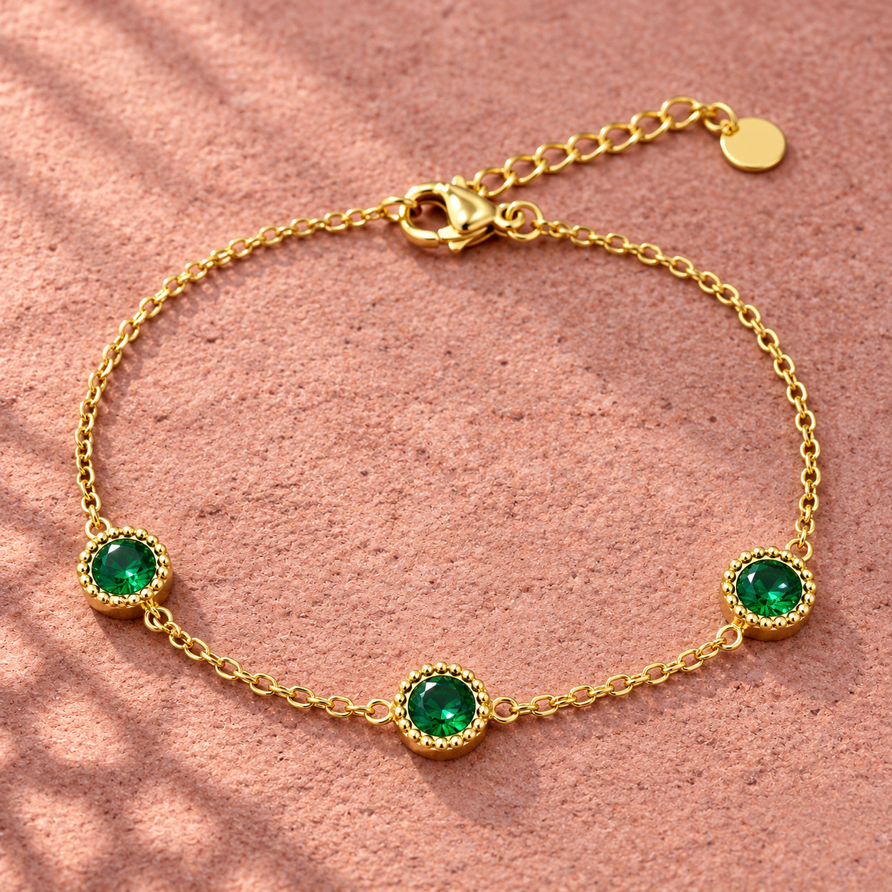 Gold-tone 316L stainless steel chain bracelet with three round green crystals in beaded bezels and a lobster clasp, laid flat on a textured surface
