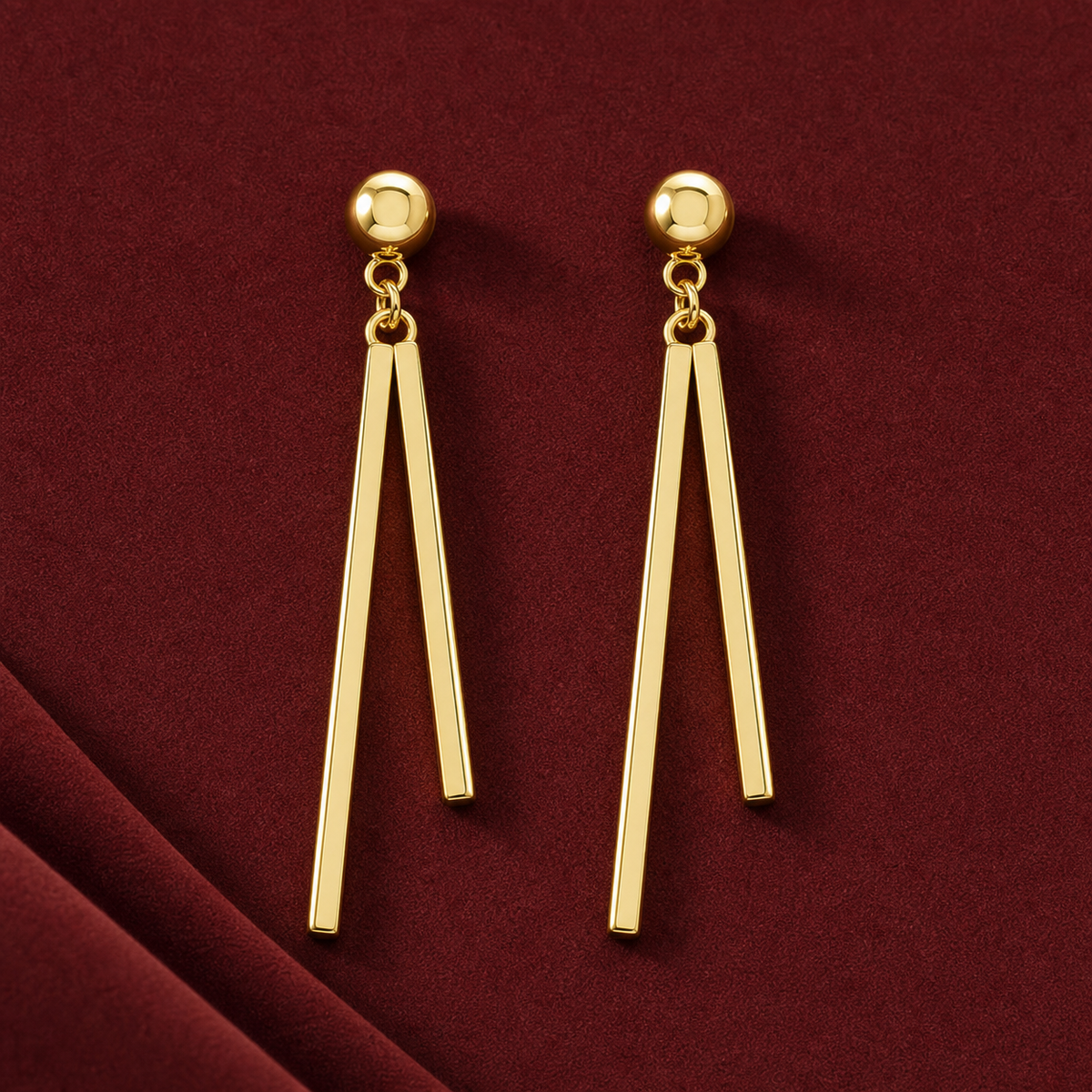 Gold-tone 304 stainless steel drop earrings with a ball stud and two long straight bars hanging from a jump ring, shown as a pair on a dark red fabric