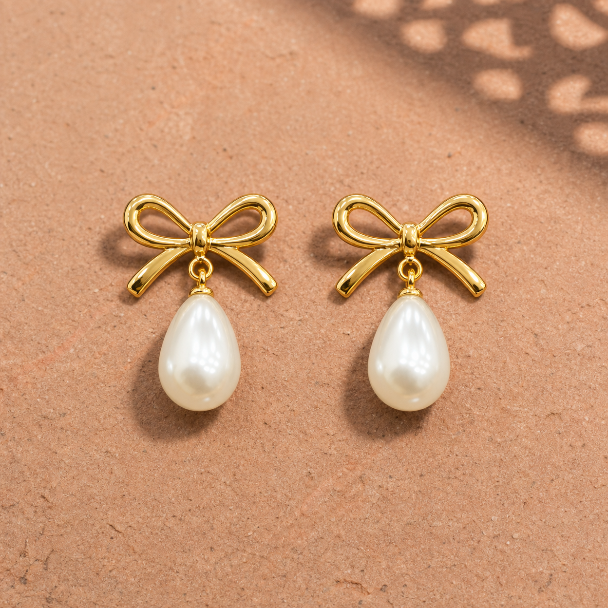 Gold-tone 304 stainless steel bow earrings with a pearl-like teardrop drop hanging from each bow, a pair shown lying flat on a sandy surface