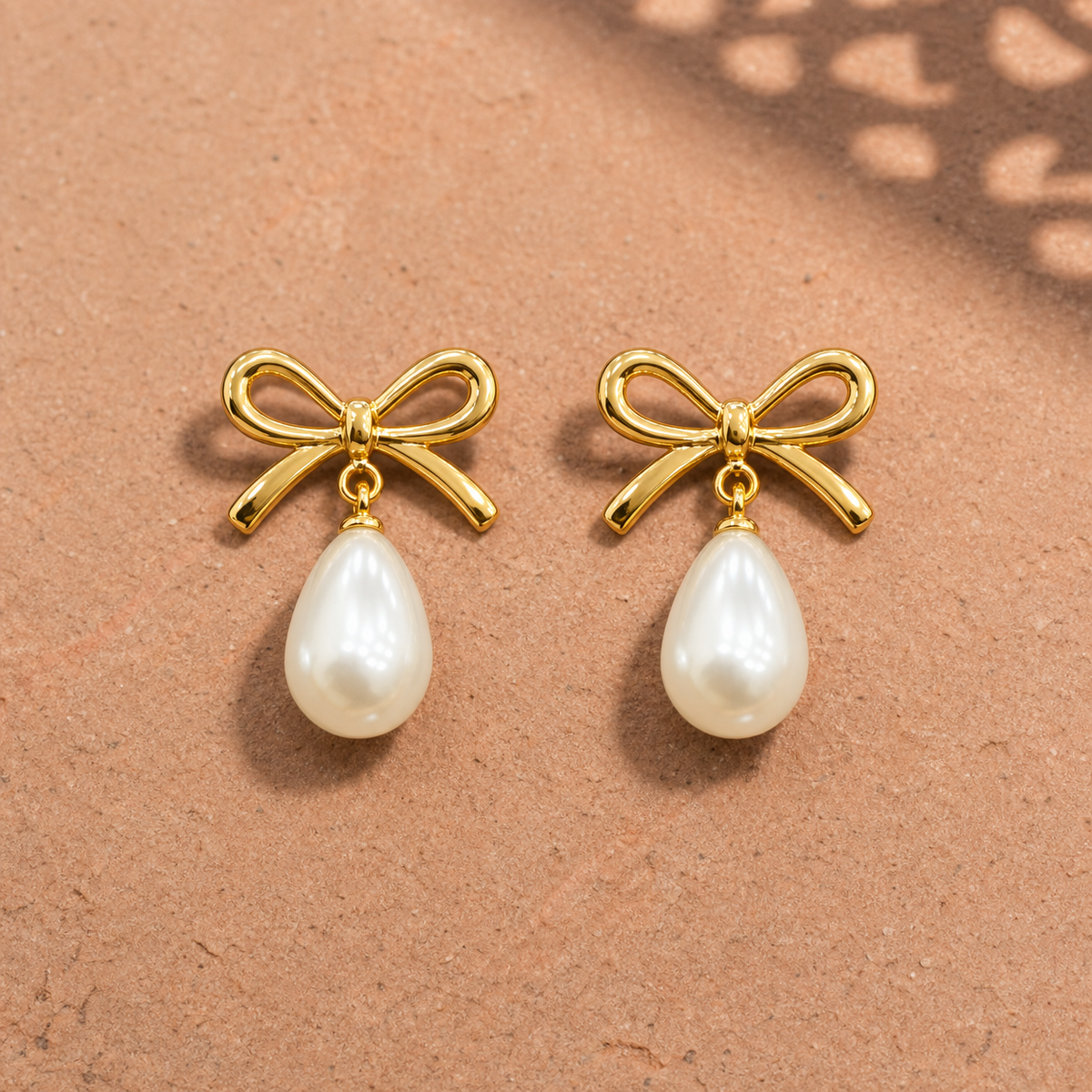 Gold-tone 304 stainless steel bow earrings with a pearl-like teardrop drop hanging from each bow, a pair shown lying flat on a sandy surface