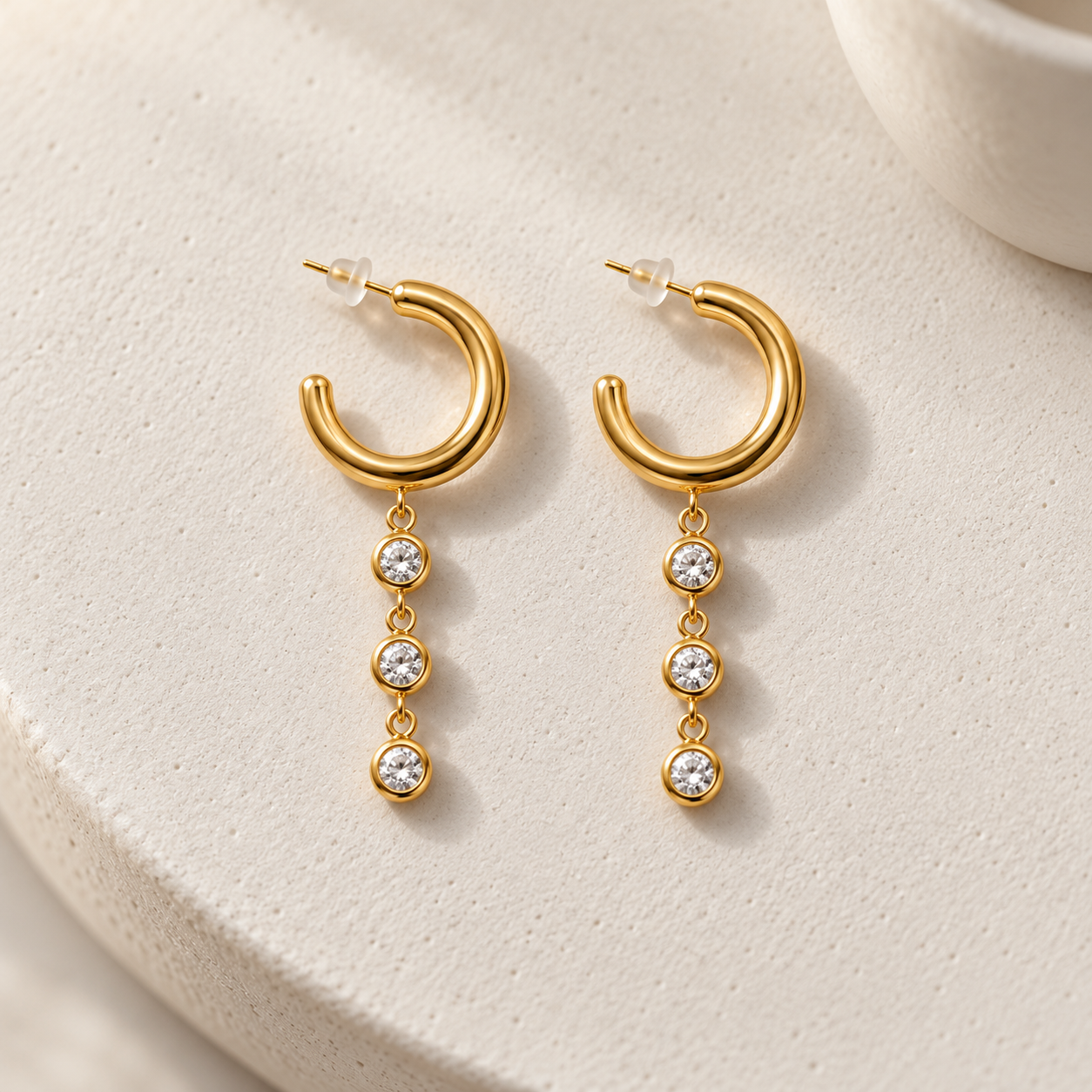 Gold-tone 316L stainless steel hoop earrings with three clear stones in bezel settings hanging from the lower edge, a pair shown on a plain background