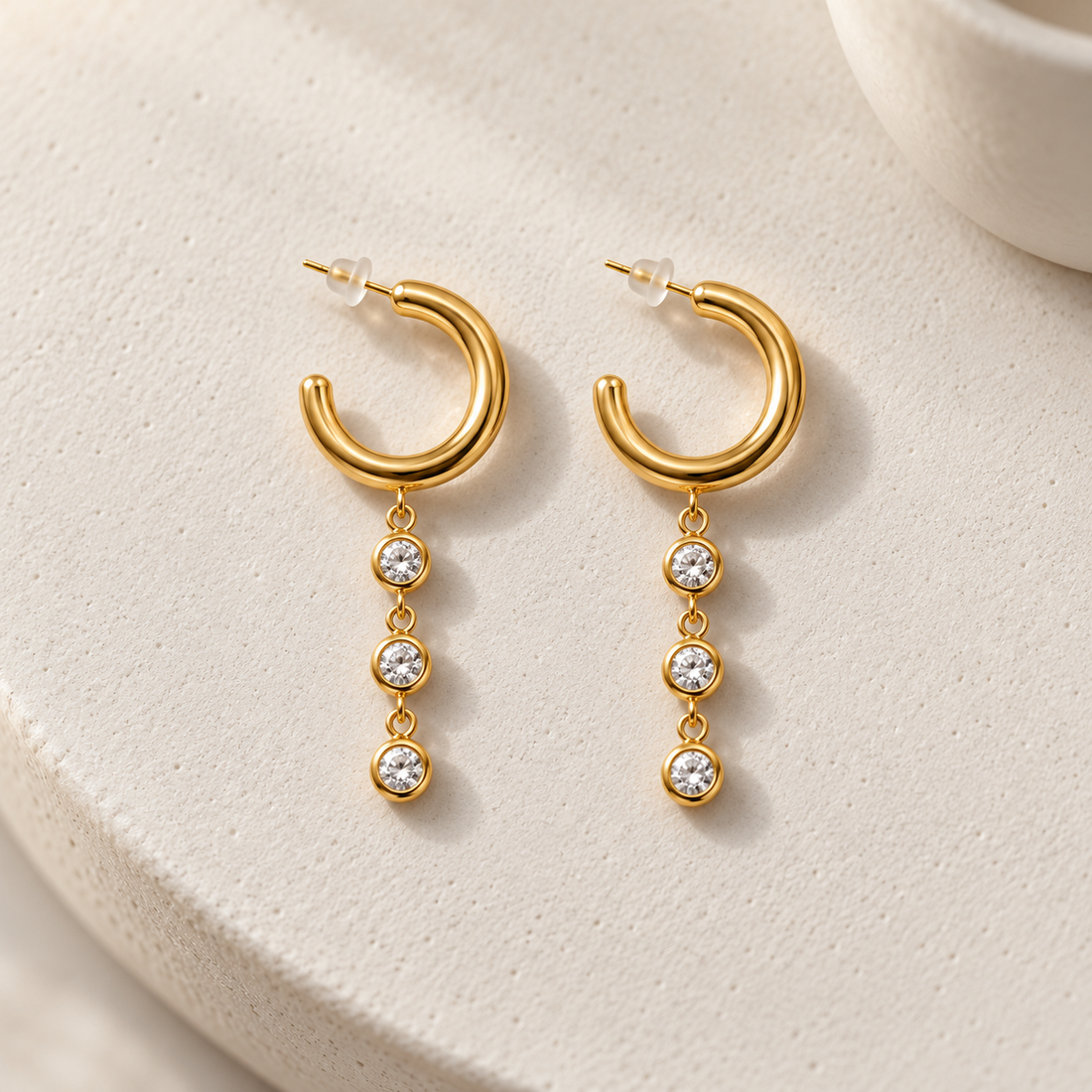 Gold-tone 316L stainless steel hoop earrings with three clear stones in bezel settings hanging from the lower edge, a pair shown on a plain background