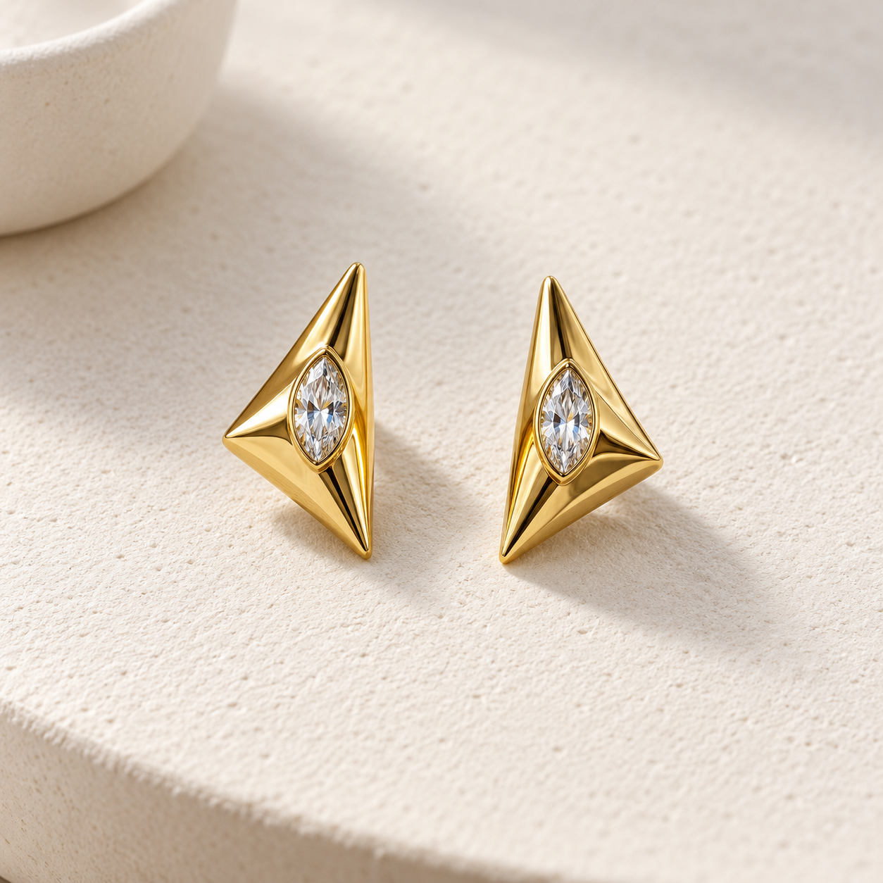 Gold-tone 316L stainless steel stud earrings with angular triangular bodies and a marquise crystal set in each, shown as a pair on a cream surface
