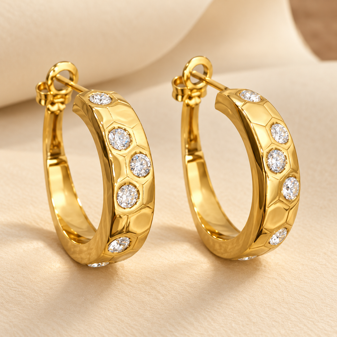 Gold-tone 316L stainless steel pair of hoop earrings with a faceted honeycomb-patterned band set with clear round crystals, shown on a cream surface