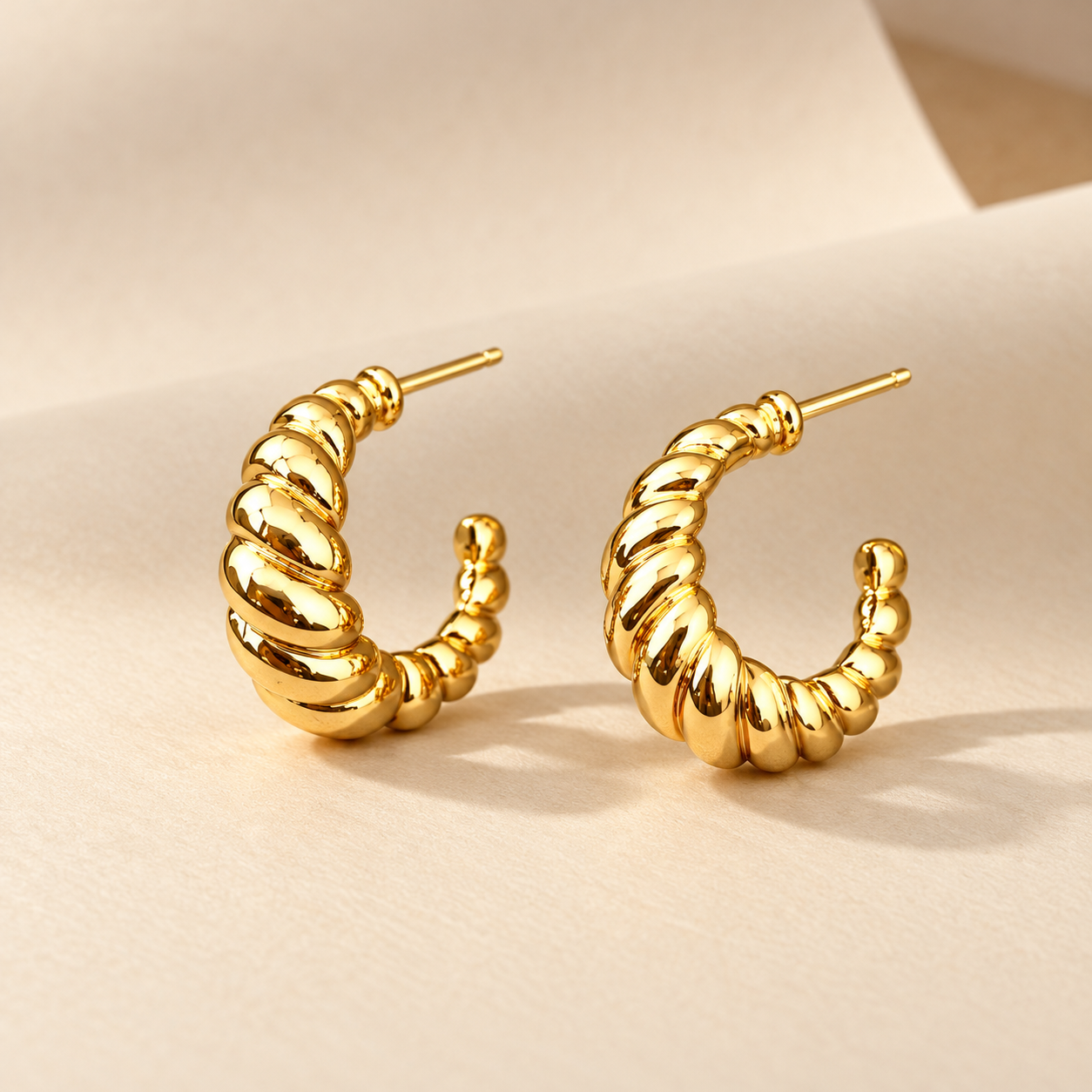 Gold-tone 304 stainless steel pair of open hoop earrings with a twisted rope texture and stud posts, shown on a cream background