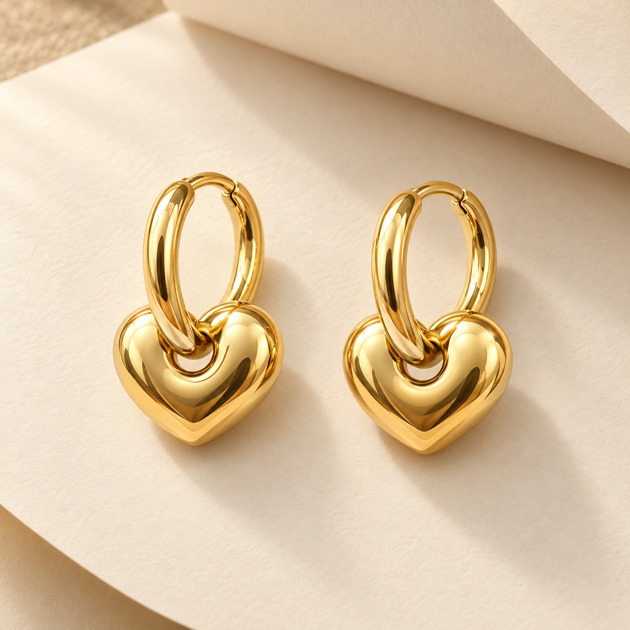 Gold-tone 316L stainless steel hoop earrings with a puffed heart charm hanging from each hoop, a pair shown flat on a cream surface