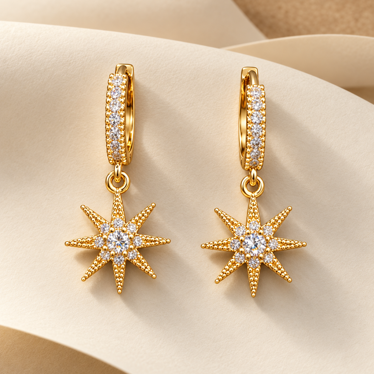Gold-tone 316L stainless steel earrings with starburst drops set with clear crystals on a pavé huggie hoop, a pair, shown on a cream backdrop