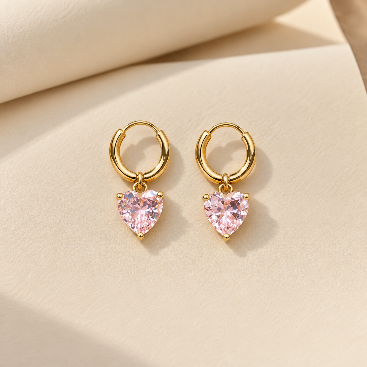 Gold-tone 316L stainless steel pair of hoop earrings with a pink heart-cut stone drop hanging from each hoop, on a cream background