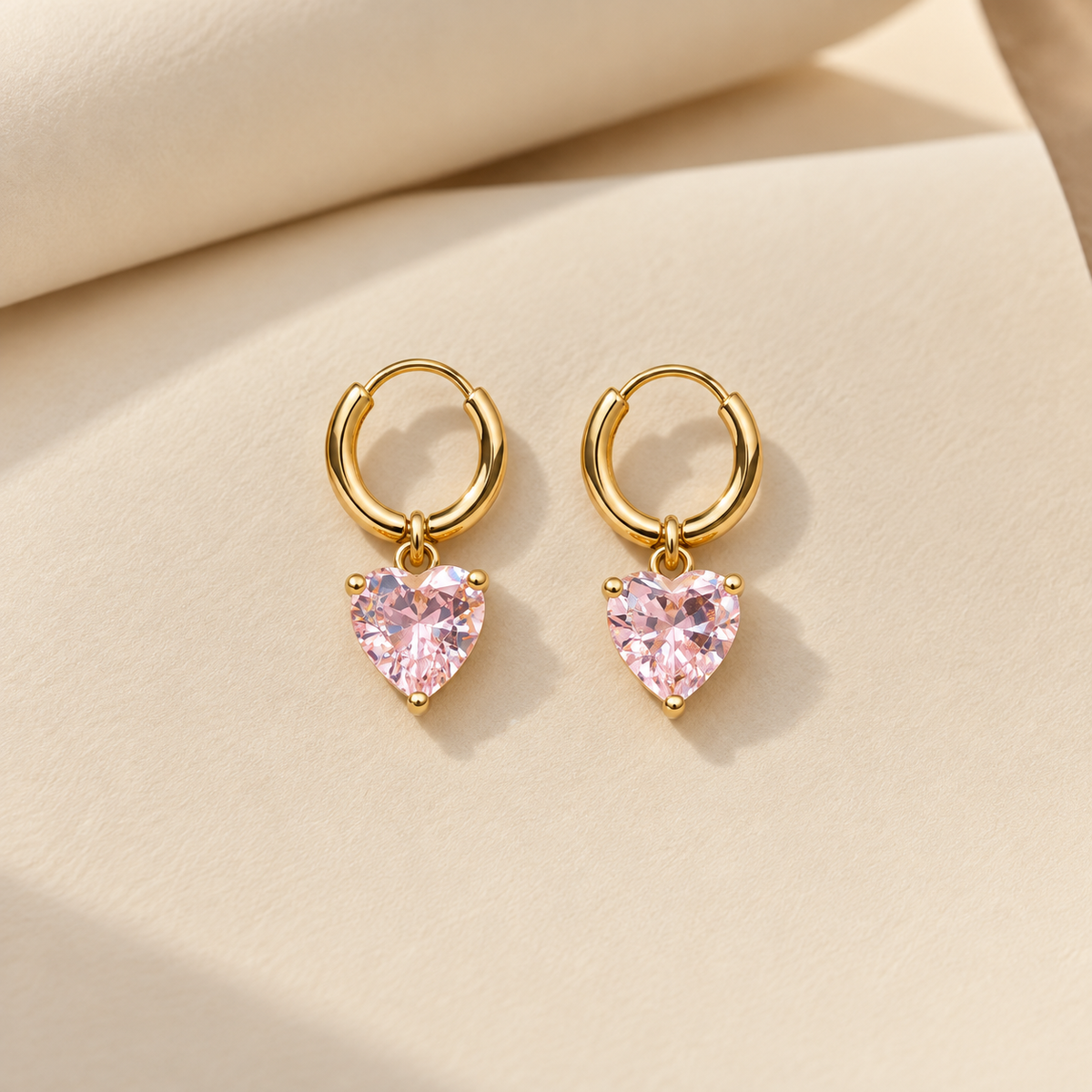 Gold-tone 316L stainless steel pair of hoop earrings with a pink heart-cut stone drop hanging from each hoop, on a cream background