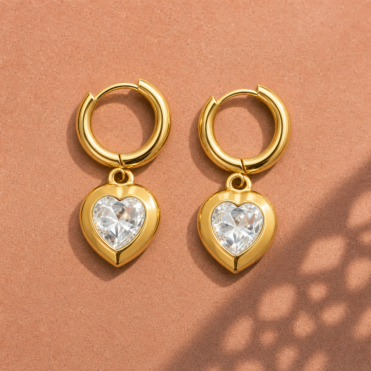 Gold-tone 304 stainless steel huggie hoop earrings with a heart-shaped clear crystal set in a bezel pendant, shown as a pair on a terracotta surface