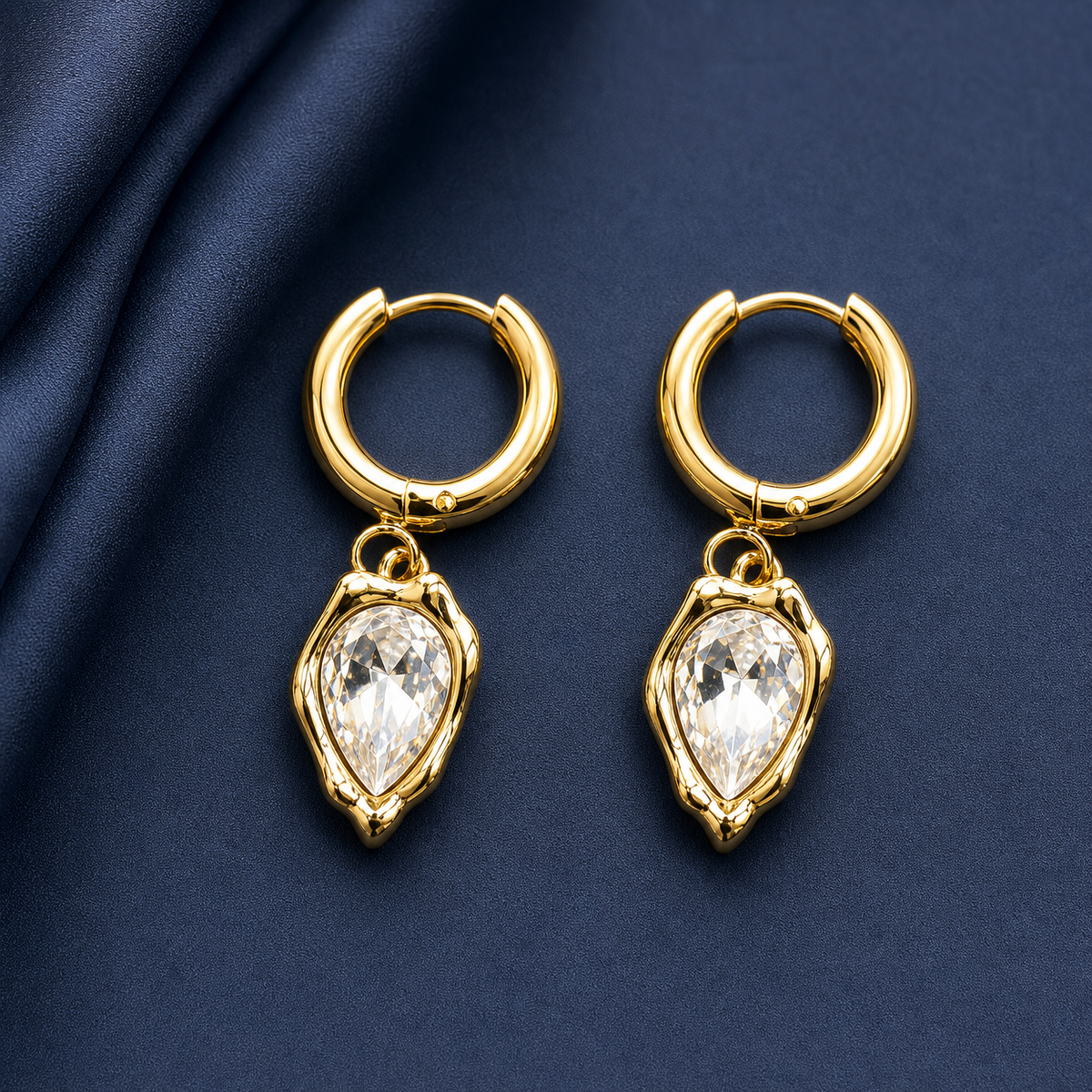 Gold-tone 304 stainless steel huggie hoop earrings with pear-cut clear crystal drops in organic gold-tone settings, a pair on a navy fabric background