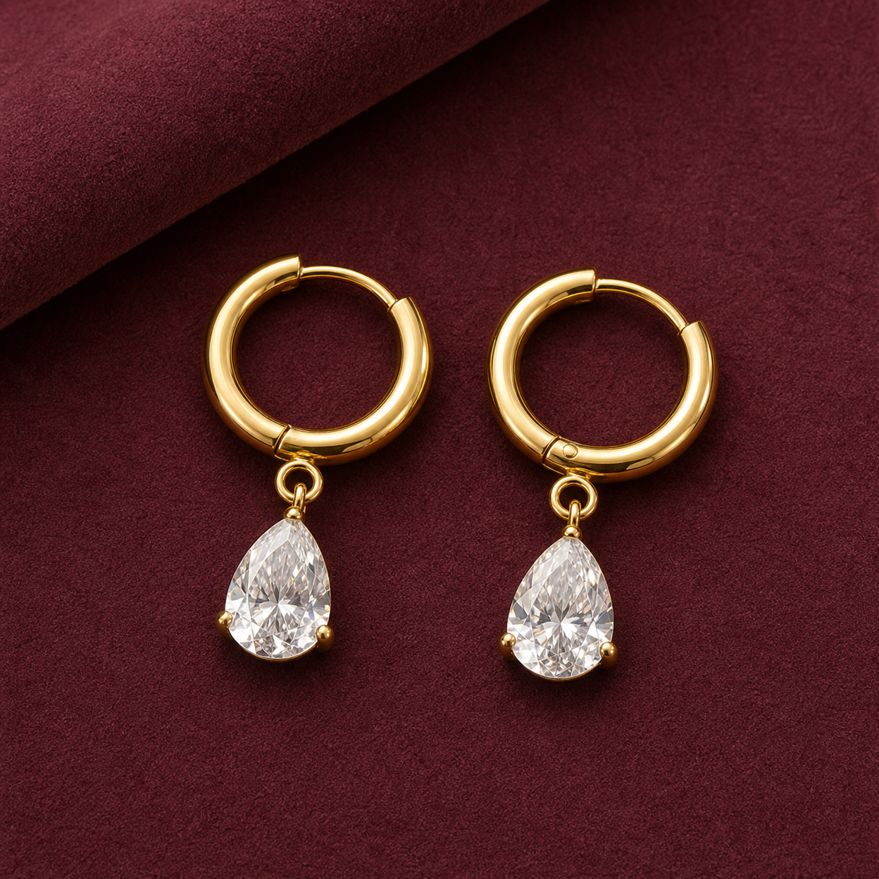 Gold-tone 316L hoop earrings with a pear-cut clear crystal drop on a pair, shown flat on a burgundy surface, a second angle