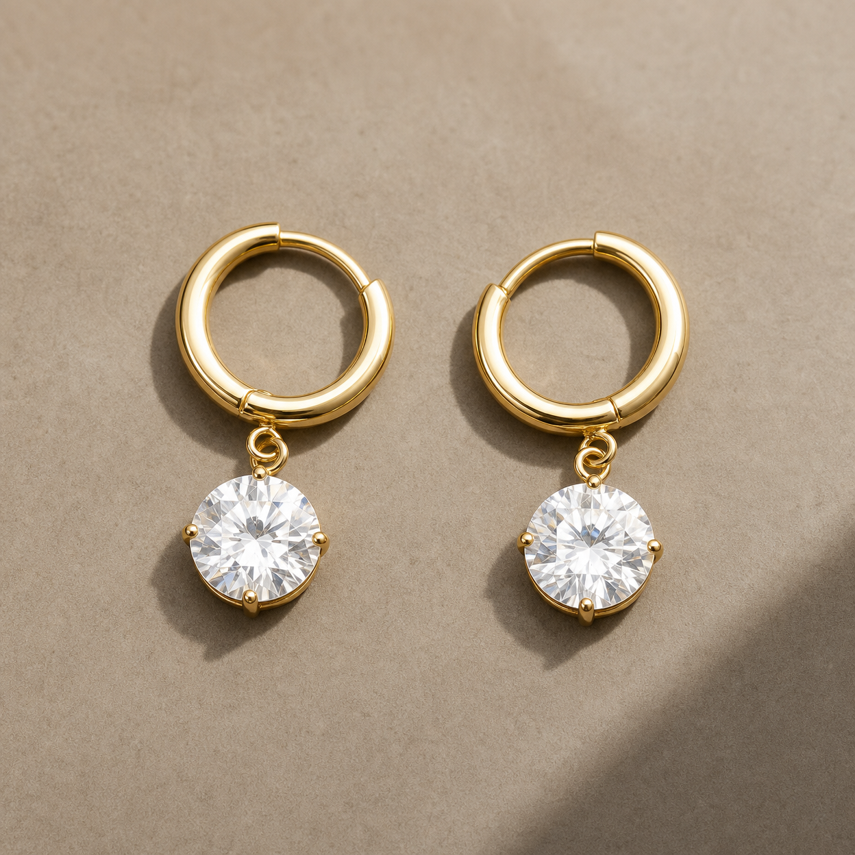 Gold-tone 316L stainless steel huggie hoop earrings with a round clear crystal drop set in four claw prongs, a pair laid flat on a beige surface