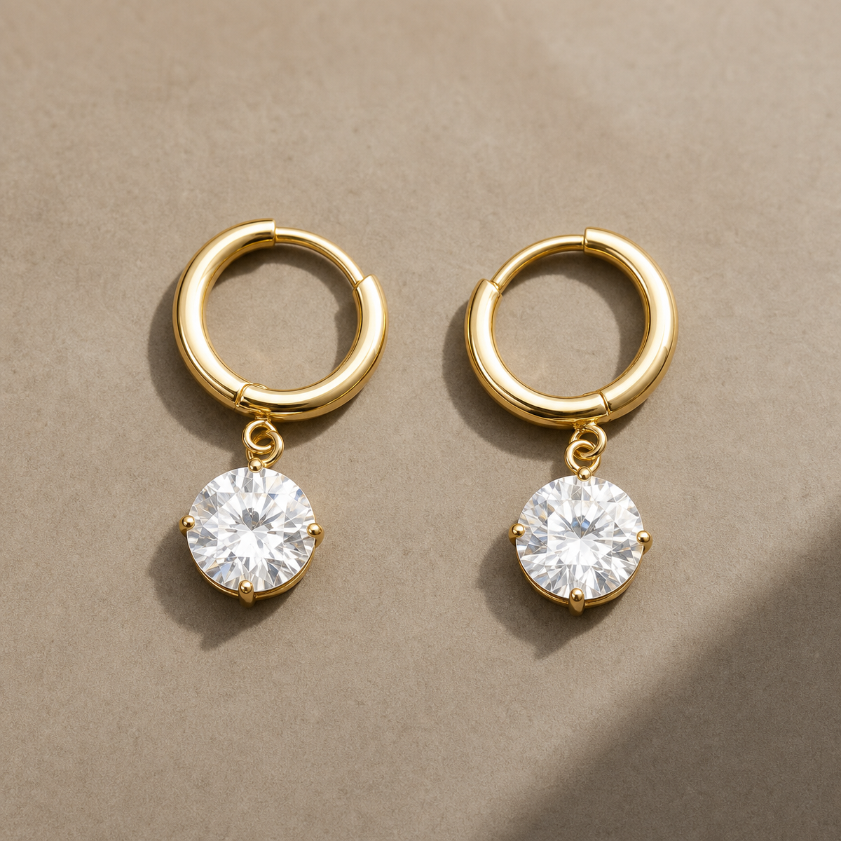 Gold-tone 316L stainless steel huggie hoop earrings with a round clear crystal drop set in four claw prongs, a pair laid flat on a beige surface