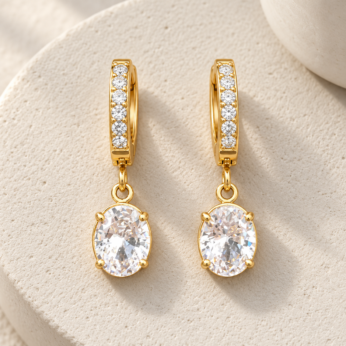 Gold-tone 316L stainless steel huggie earrings with clear crystal-set hoops and an oval crystal drop, a pair shown on a stone surface