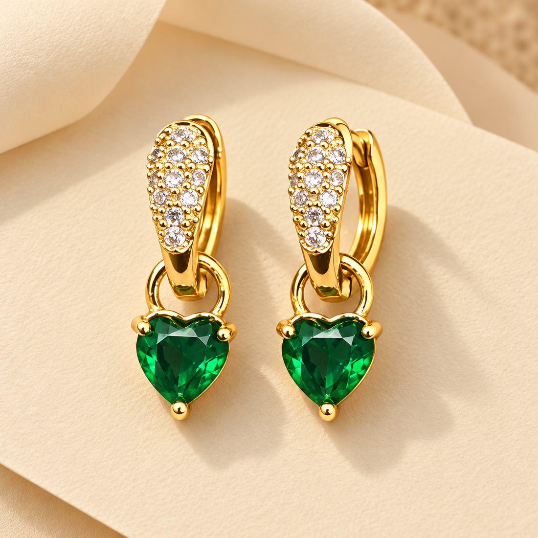 Gold-tone 316L stainless steel pair of huggie hoop earrings with pavé clear stones and a dangling green heart-cut gem drop, shown on a cream fabric background