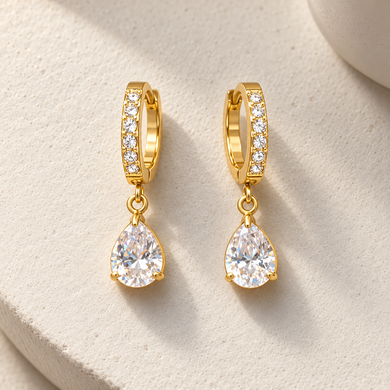 Gold-tone 316L stainless steel huggie earrings with a row of clear stones and a pear-cut clear stone drop, a pair on a cream surface