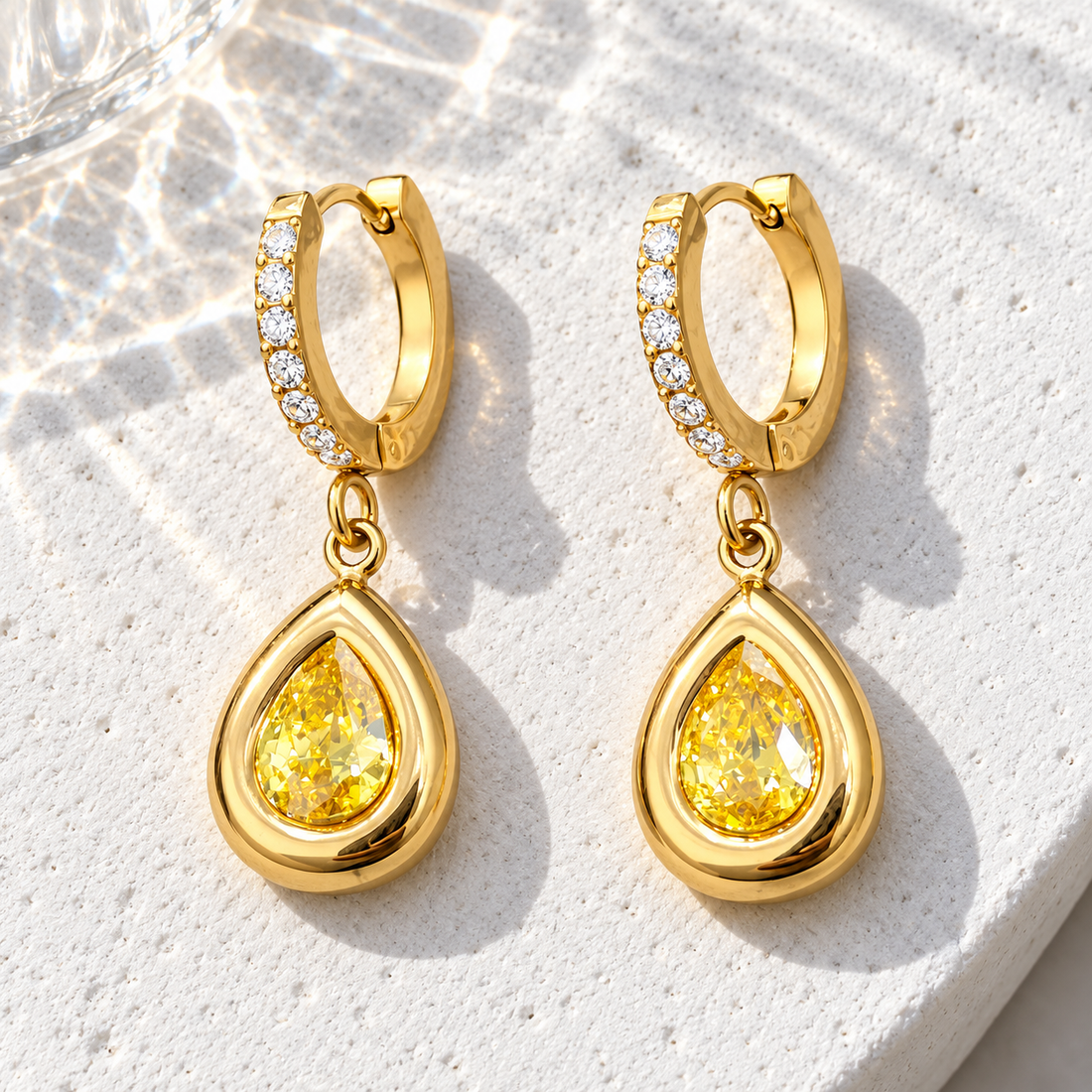 Gold-tone 316L stainless steel pair of huggie hoop earrings with clear stones and a yellow pear-cut drop, on a white stone surface