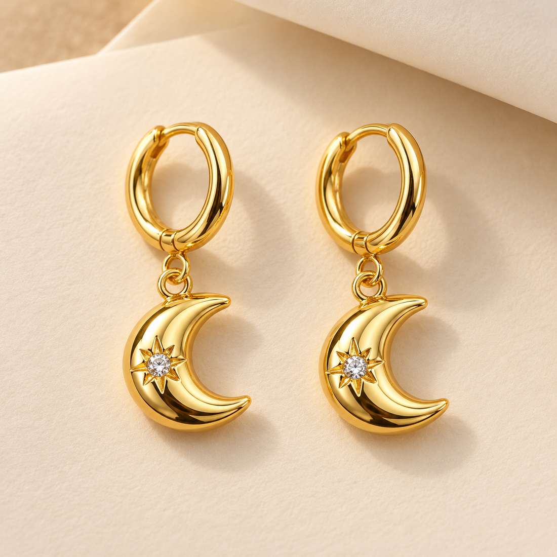 Gold-tone 316L stainless steel huggie earrings with crescent moon drops set with a clear stone in a star motif, shown as a pair on a cream background