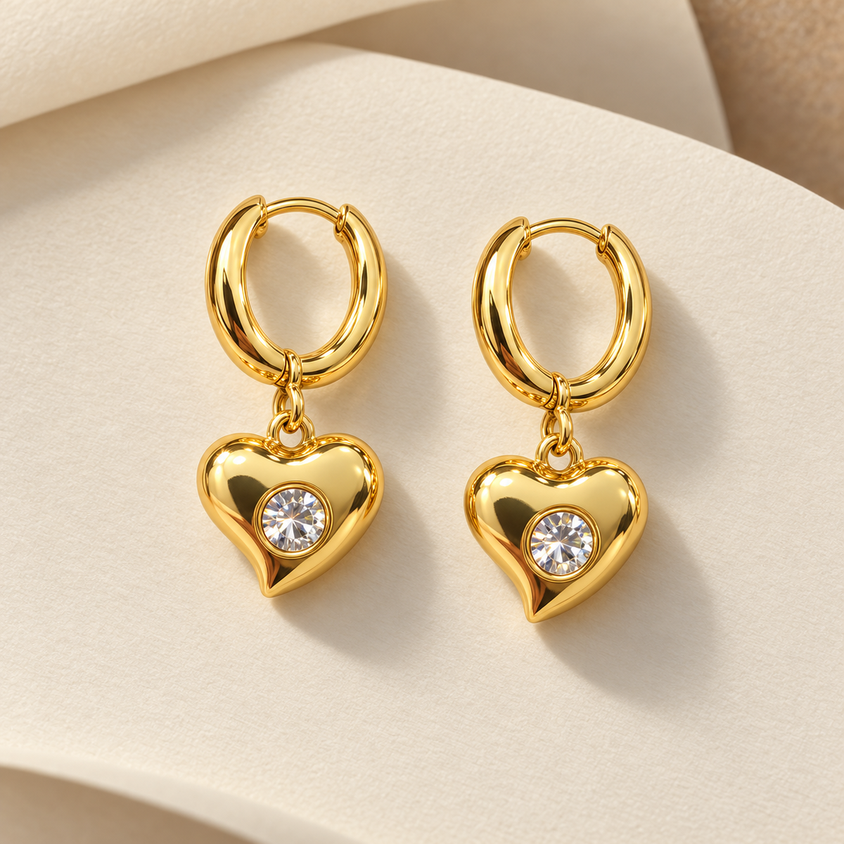Gold-tone 316L stainless steel huggie hoop earrings with puffed heart drops set with a round clear crystal, a pair on a cream surface