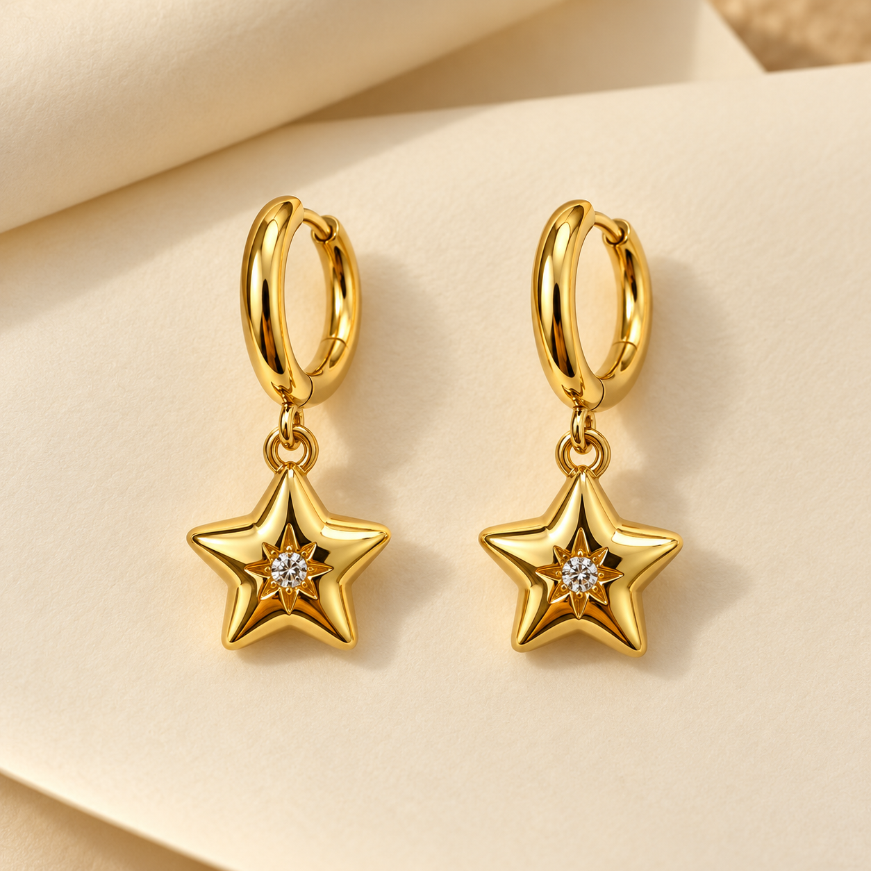 Gold-tone 316L stainless steel hoop earrings with puffed star drops set with a clear stone at the centre, shown as a pair on a cream background