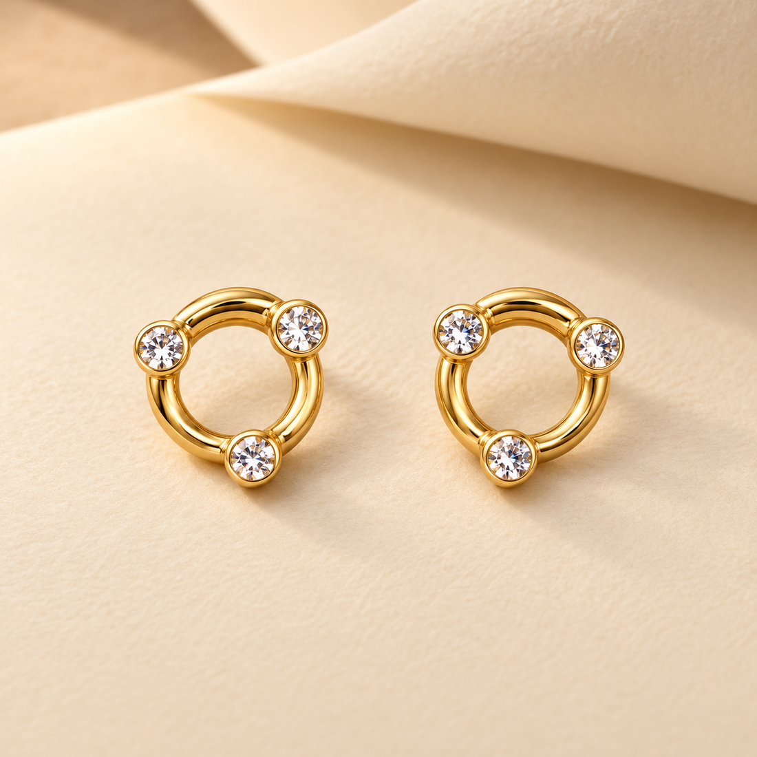 Gold-tone 316L stainless steel earrings, a pair of open circle hoops with three bezel-set clear stones spaced around each ring, shown on a cream surface