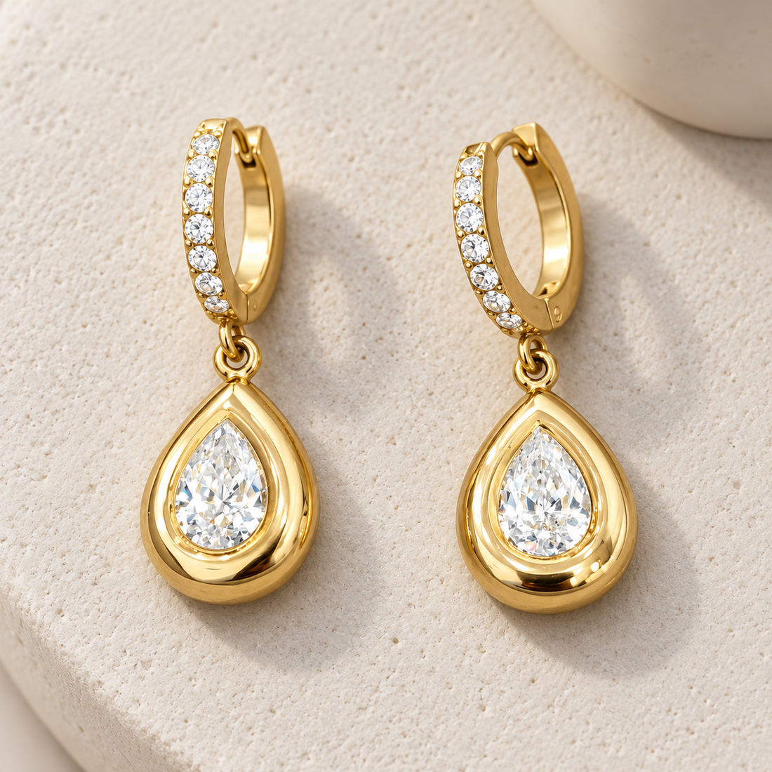 Gold-tone 316L stainless steel huggie hoop earrings with crystal-set hoops and a pear-shaped clear stone drop, a pair on a stone surface