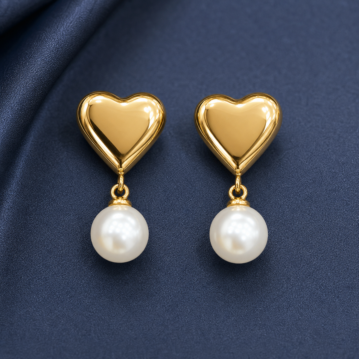 Gold-tone 304 stainless steel drop earrings with a polished heart stud and a hanging white pearl bead, shown as a pair on navy fabric