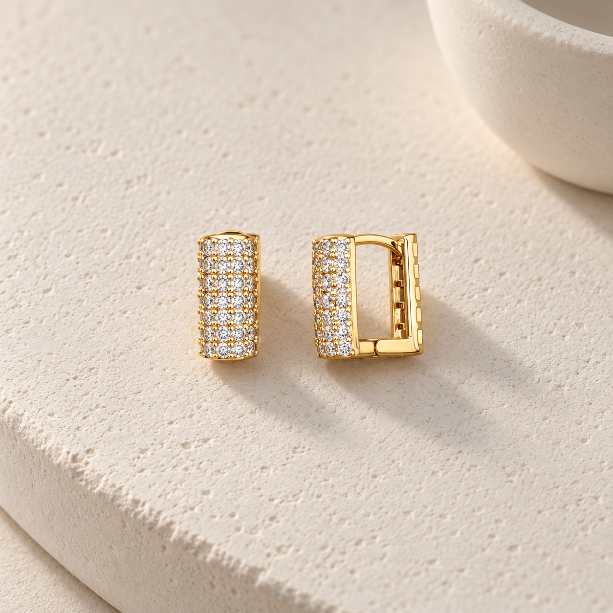 Gold-tone 316L stainless steel huggie earrings with rows of clear crystals across a square-profile band, shown as a pair on a stone surface