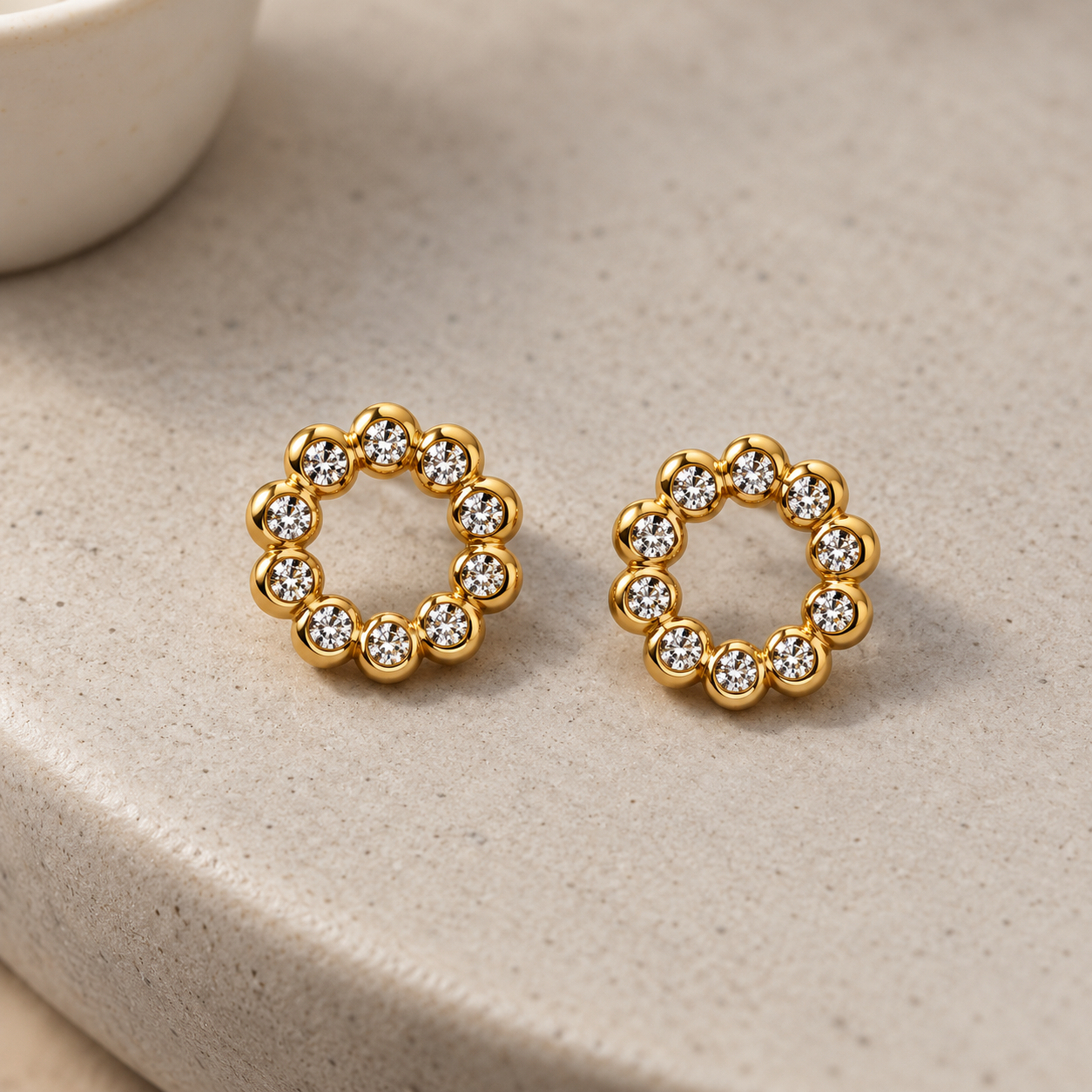 Gold-tone 316L stainless steel stud earrings in an open circle of clear stones in bezel settings, a pair on a stone surface