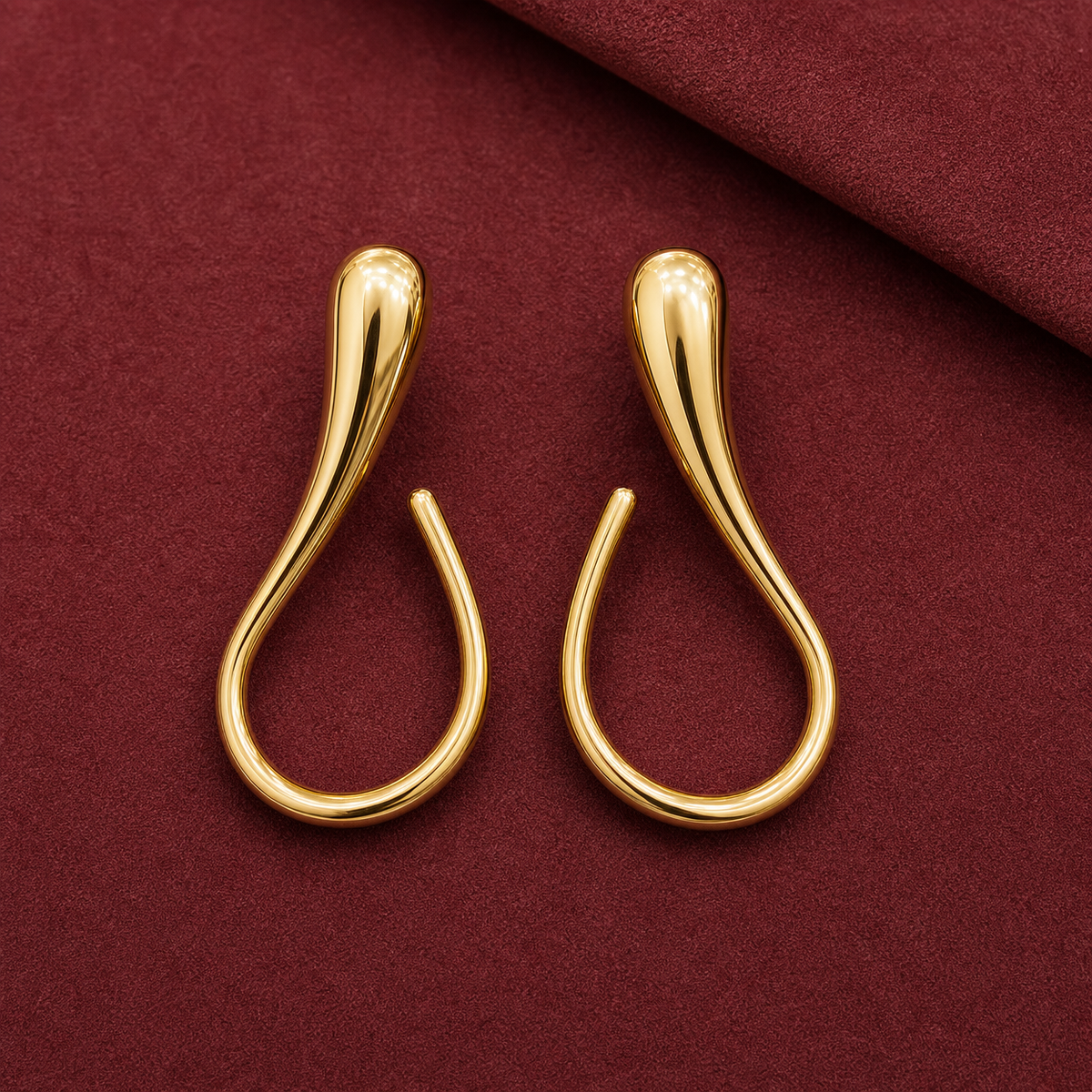 Gold-tone 304 stainless steel drop earrings in a teardrop loop shape with a smooth polished finish, shown as a pair on a burgundy surface