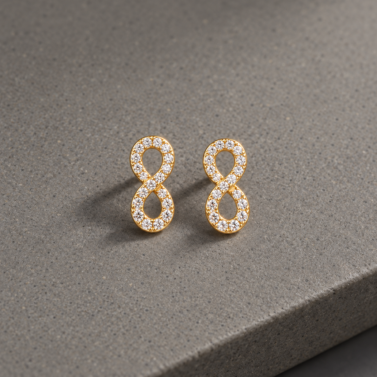 Gold-tone 316L stainless steel infinity-shaped stud earrings set with clear crystals, shown as a pair on a grey stone surface