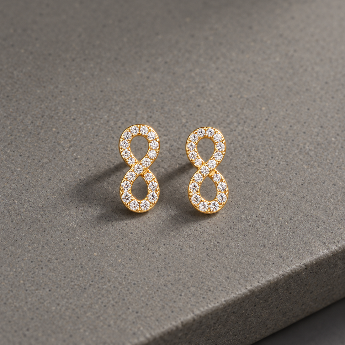 Gold-tone 316L stainless steel infinity-shaped stud earrings set with clear crystals, shown as a pair on a grey stone surface