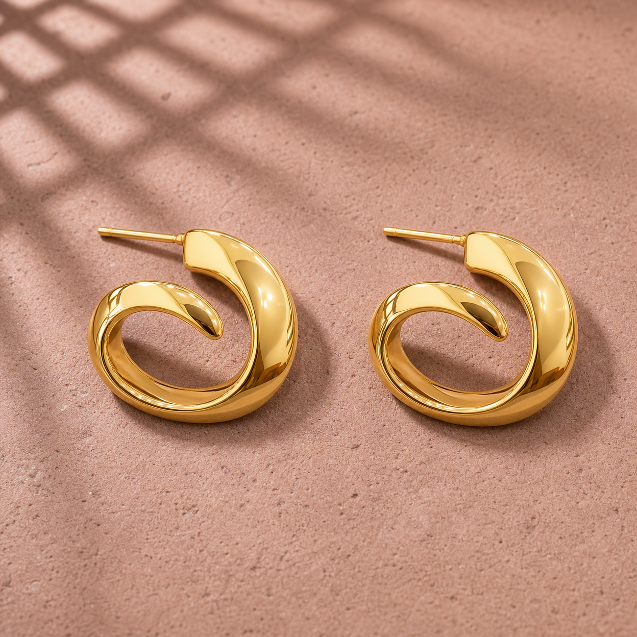 Gold-tone 304 stainless steel pair of swirl hoop earrings with a thick tapering spiral band and a post back, shown laid flat on a pink surface