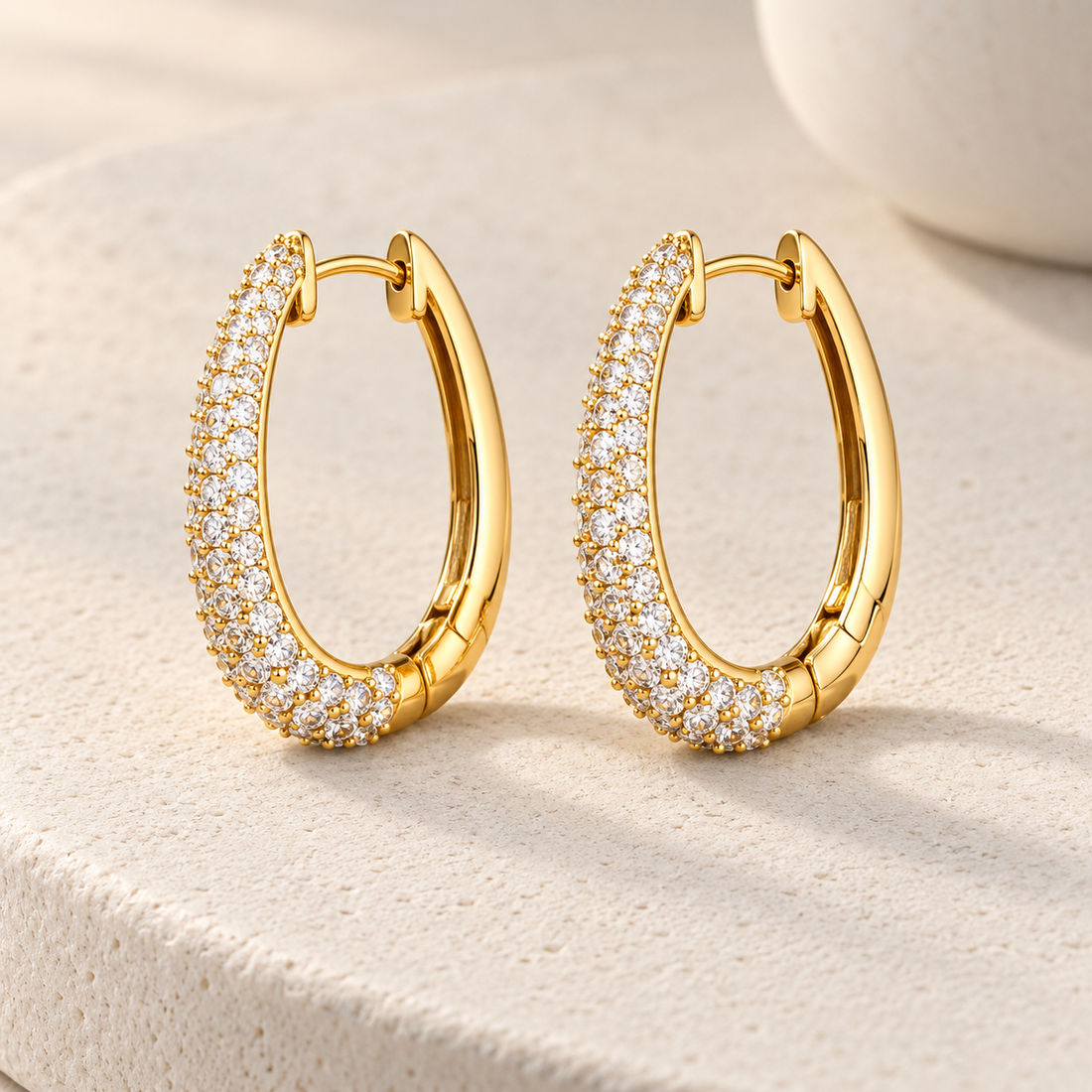 Gold-tone 316L stainless steel oval hoop earrings with a pavé band of clear crystals, a pair shown on a stone surface