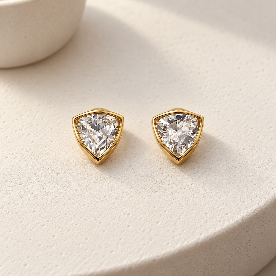 Gold-tone 316L stainless steel stud earrings with trillion-cut clear stones in bezel settings, shown as a pair on a cream surface