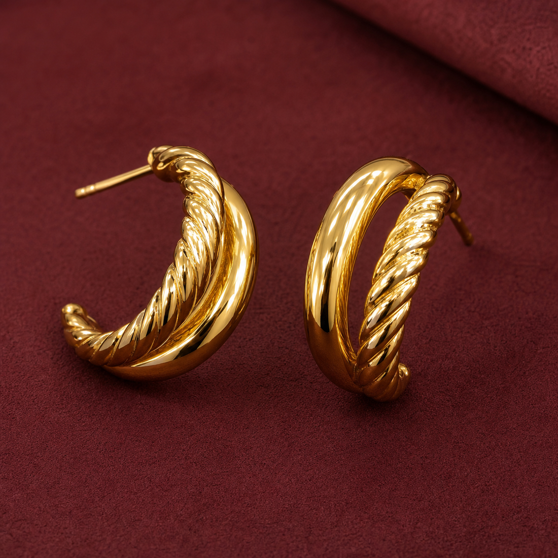 Gold-tone 304 stainless steel pair of hoop earrings with a smooth band crossed over a twisted rope band, with post backs, shown on a maroon surface
