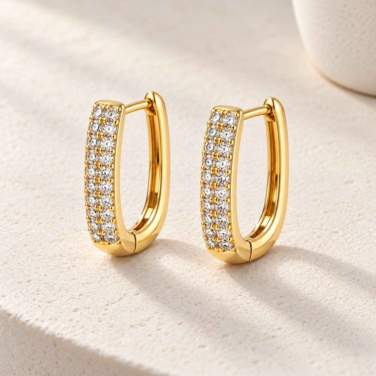 Gold-tone 316L stainless steel pair of U-shaped huggie hoop earrings with two rows of clear crystals along the front, shown on a cream surface