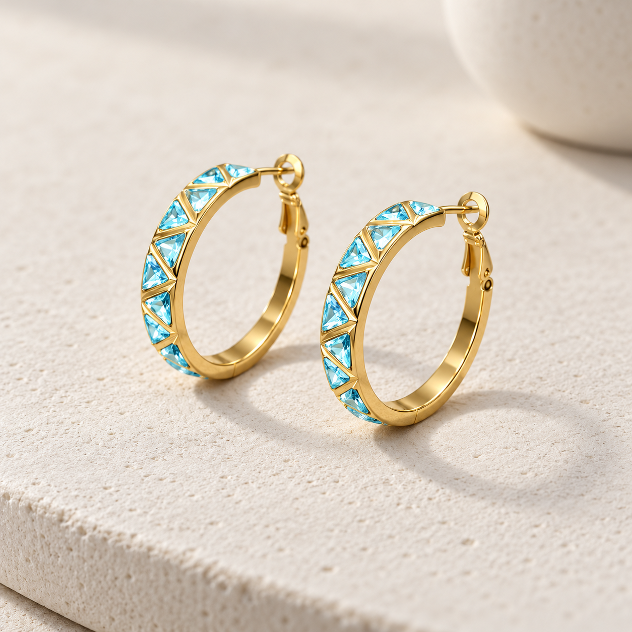 Gold-tone 316L stainless steel hoop earrings with a row of aqua triangular stones set along the front, a pair shown on a light stone surface