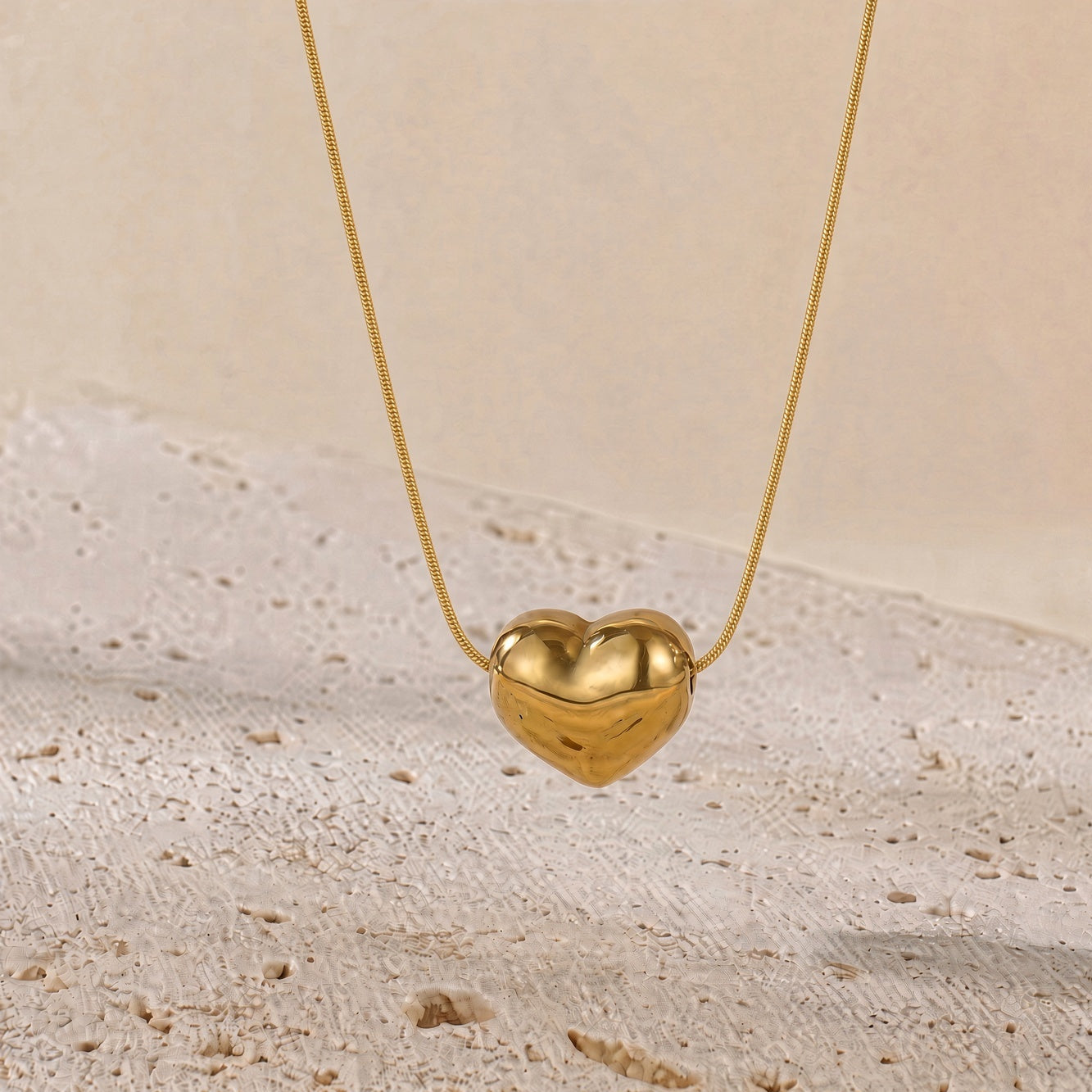 A gold heart-shaped pendant necklace hanging from a delicate gold chain against a warm beige textured background.