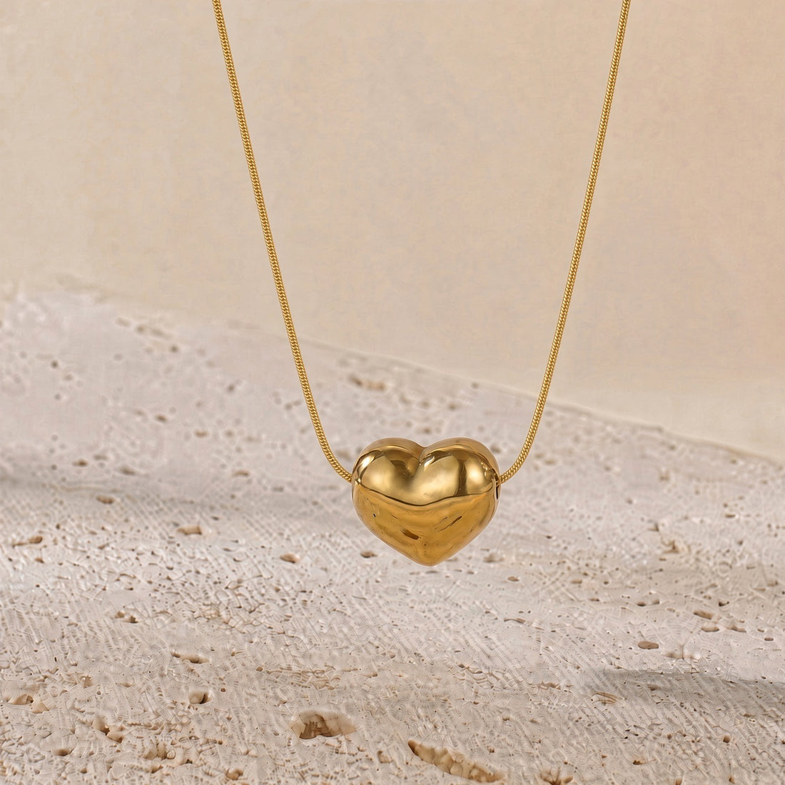 A gold heart-shaped pendant necklace hanging from a delicate gold chain against a warm beige textured background.