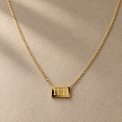 Gold-tone 316L stainless steel necklace with a fine cable chain and a rectangular bar pendant engraved with 11:11, laid flat on a beige surface