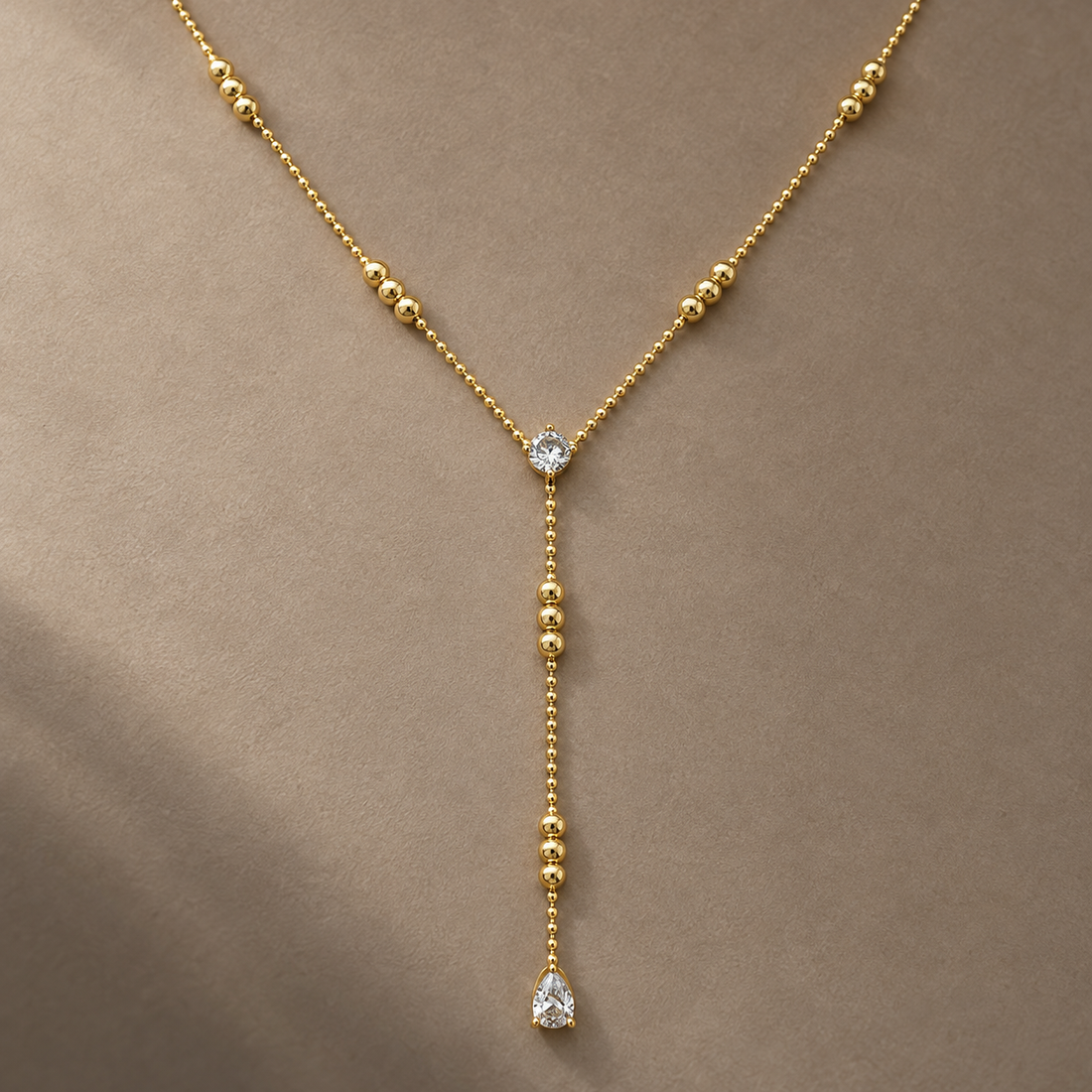 Gold-tone 316L stainless steel lariat necklace with a ball chain, graduated gold beads and a round and pear-cut clear stone drop, worn on a neck