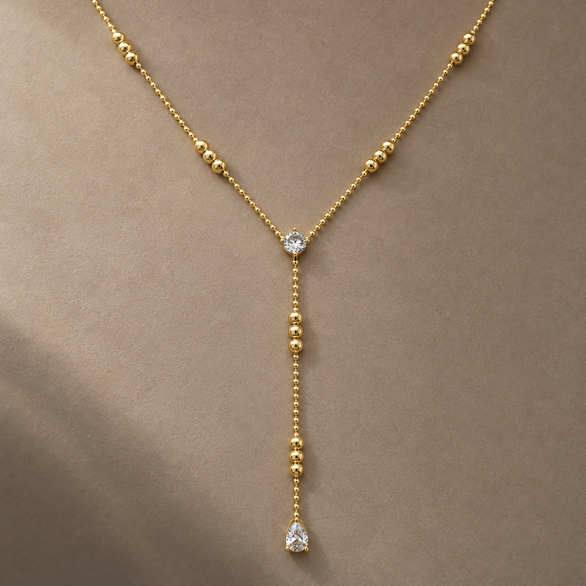 Gold-tone 316L stainless steel lariat necklace with a ball chain, graduated gold beads and a round and pear-cut clear stone drop, worn on a neck