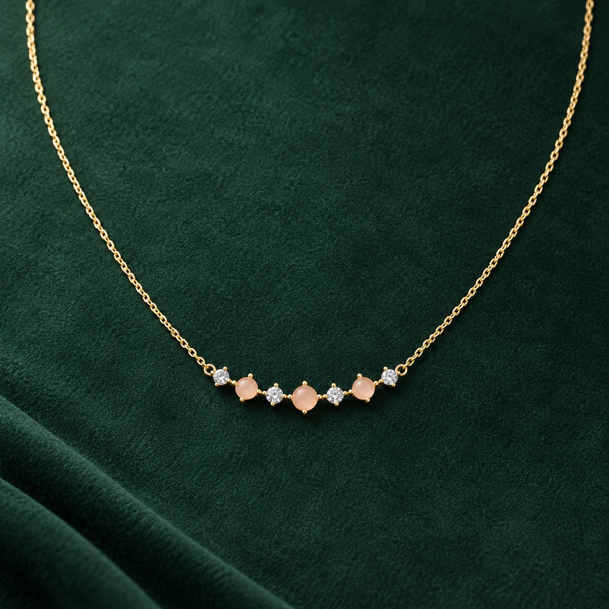 Gold-tone 316L necklace with a fine cable chain and a curved bar of peach stones alternating with clear crystals, laid flat on green fabric