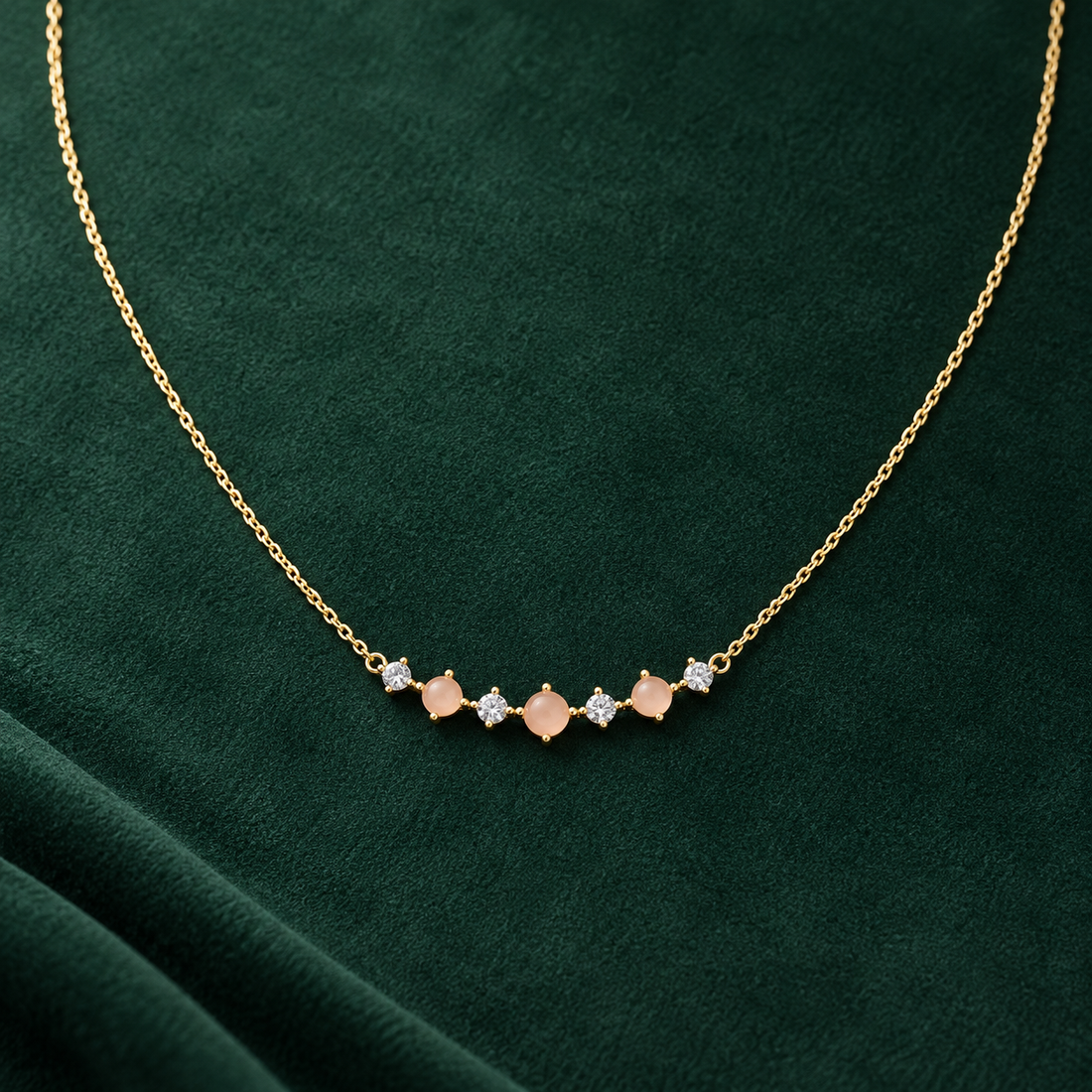 Gold-tone 316L necklace with a fine cable chain and a curved bar of peach stones alternating with clear crystals, laid flat on green fabric