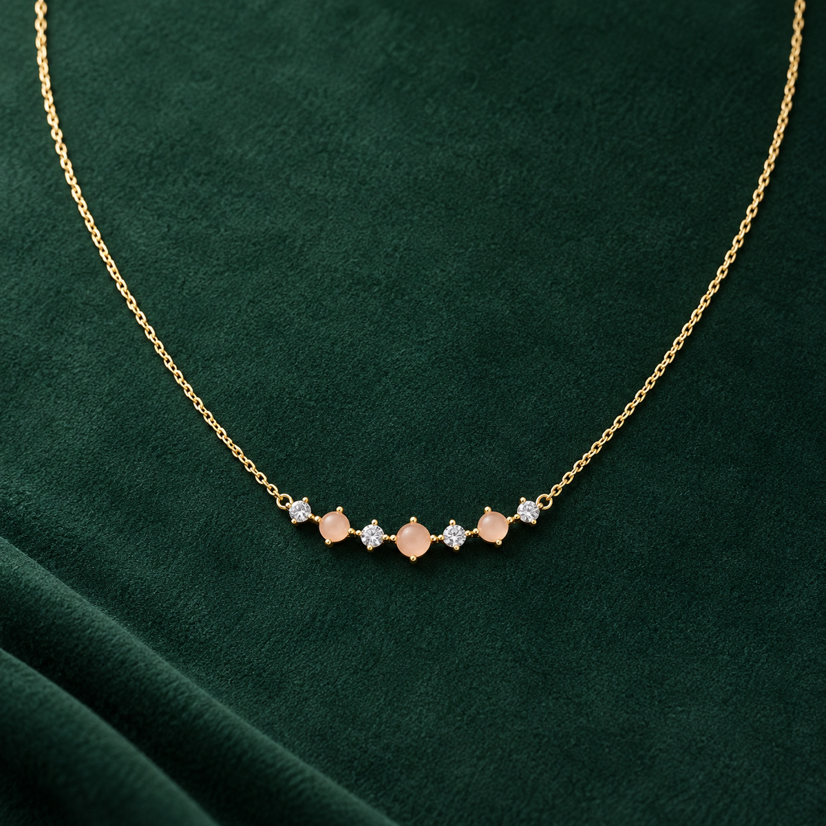 Gold-tone 316L necklace with a fine cable chain and a curved bar of peach stones alternating with clear crystals, laid flat on green fabric