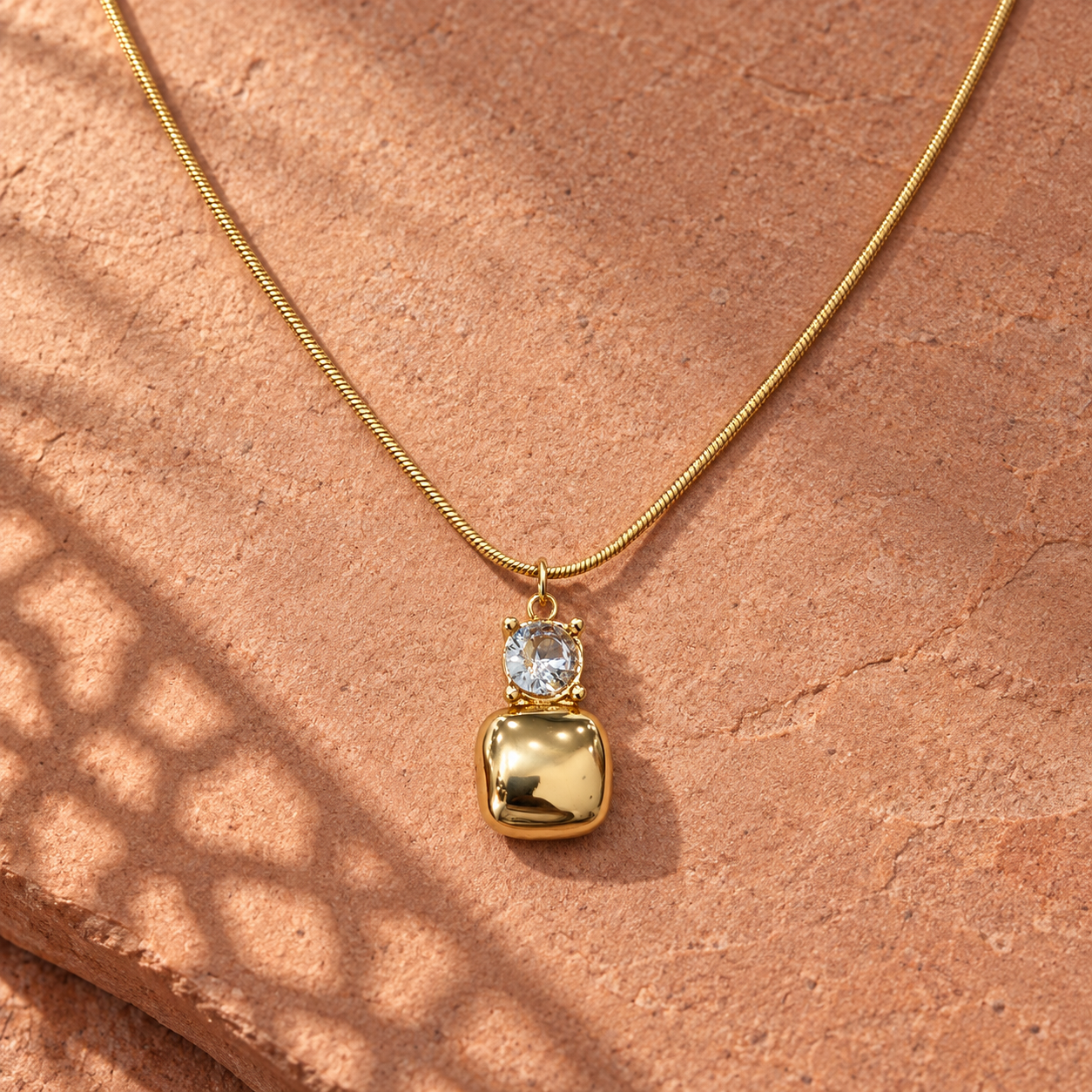 Gold-tone 316L snake chain necklace with a round clear crystal and a puffy cushion-shaped pendant, shown laid flat on stone