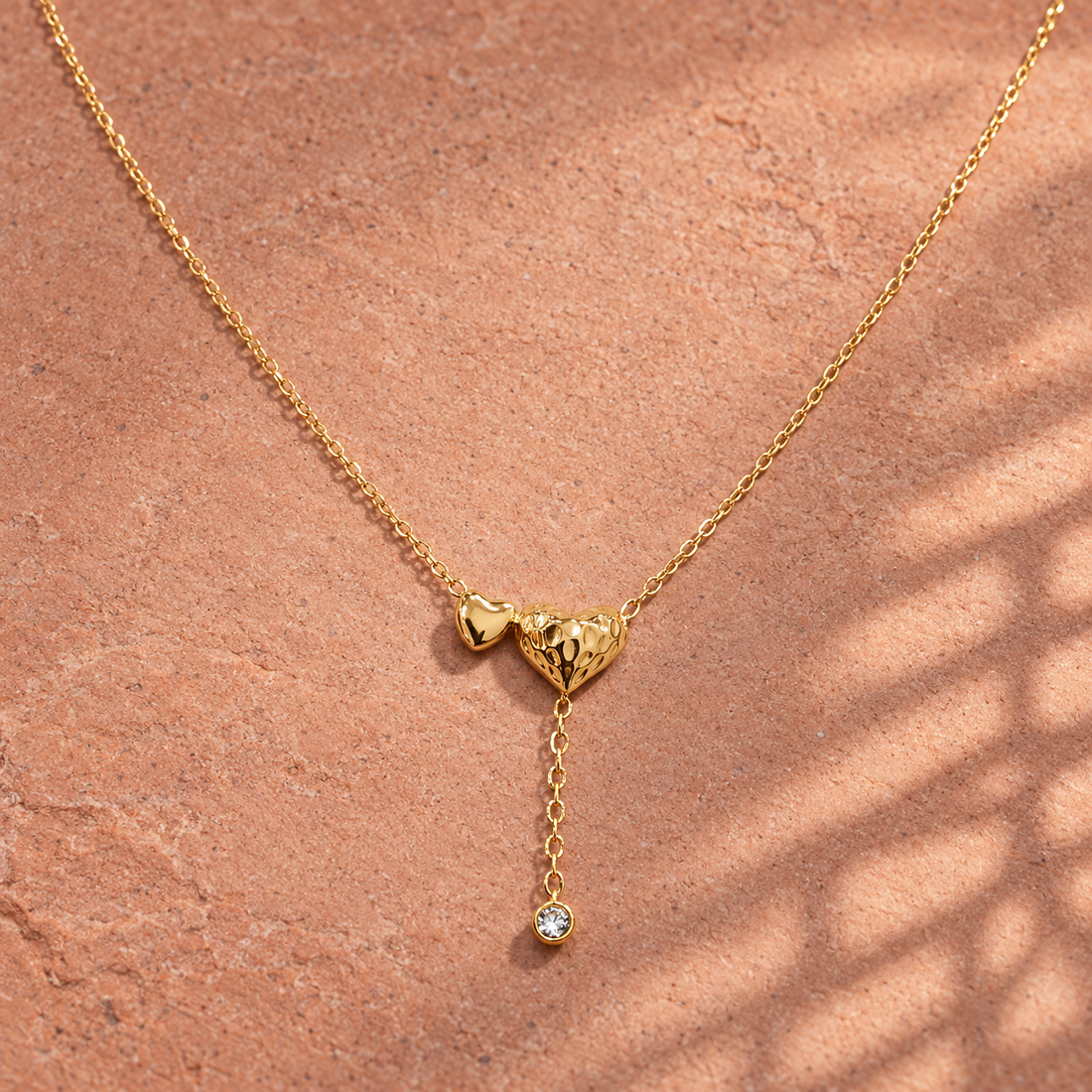 Gold-tone 316L stainless steel necklace with two heart pendants, one textured, and a drop chain ending in a clear crystal, lying on a stone surface